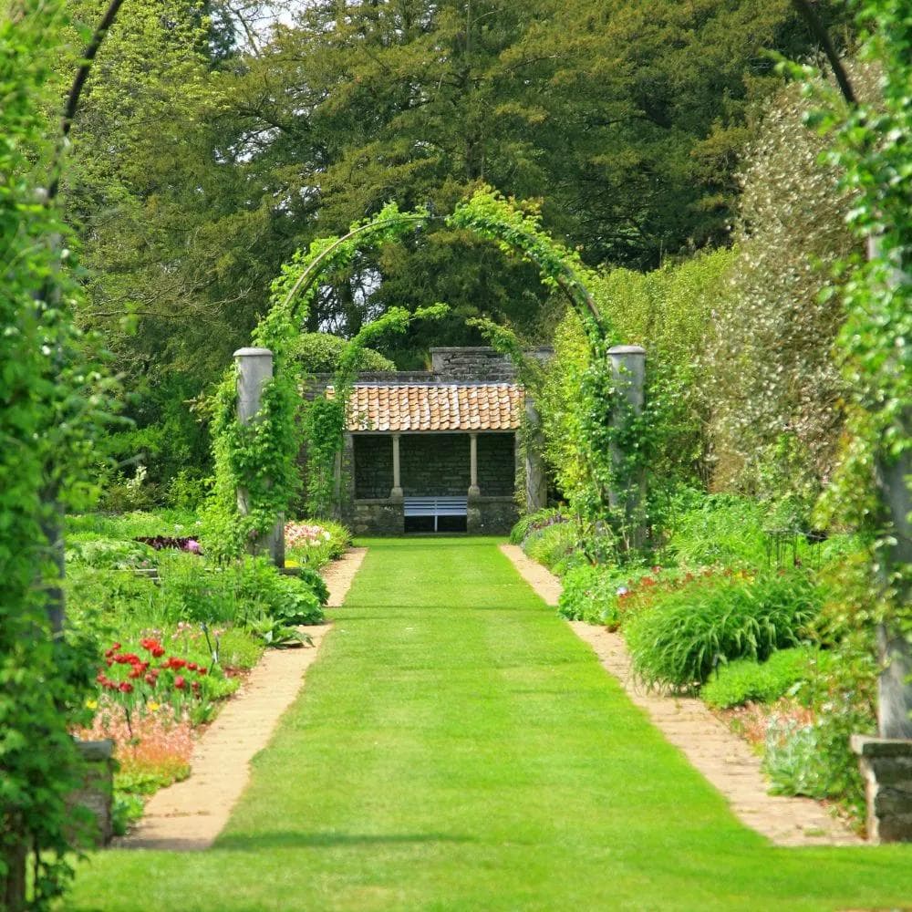 Garden Stewardship - Long, narrow garden flanked by bushes and colourful flowers ending with a small bench and awning